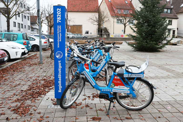 Es wird das Regiorad Station gezeigt. Dies steht an einer blauen Säule. Das Fahrrad selbst ist auch blau. Es steht auf einem gepflasterten Boden. Links sind einige Autos zu sehen. 
